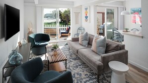 A modern living room with a sofa, two armchairs, a coffee table, and a TV mounted on the wall. There is a sliding glass door leading to an outdoor area with a view of palm trees. - Waterfront Condo w/ AC, BBQ, & Outdoor Living (San Diego)