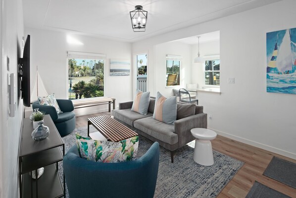 Una sala de estar moderna con un sofá gris, un sillón azul y una mesa de centro de madera. Hay un inodoro blanco y una mesita con un jarrón. Amplias ventanas ofrecen vistas a palmeras y a una playa.
