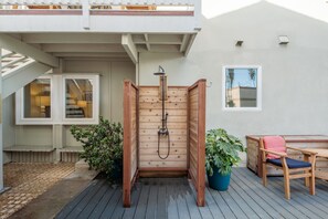Ducha exterior de madera rodeada por un cerco de madera, una terraza, una silla y plantas en macetas.