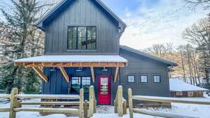 A two-story wooden cabin with a red door, a covered porch, and a fenced front yard. - New Construction Lakefront Home with Private Dock, Fire Pit, Hot Tub (Swanton)