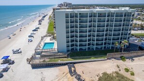 Un hôtel à plusieurs étages doté d’une piscine et d’un accès à la plage, situé près d’une plage où l’on peut voir des voitures garées et des parasols.