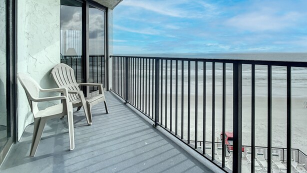 Un balcon avec deux chaises blanches, une porte vitrée et une vue sur la plage.