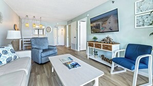 A modern living room with a sofa, armchair, and coffee table. There is a TV mounted on the wall, a lamp, and a decorative wall piece.