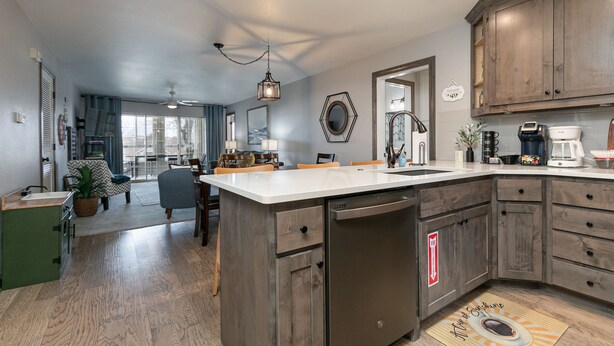 A modern kitchen with wooden cabinets, a dishwasher, and a large island. There is a dining area with chairs and a table, and a view of the outdoors through large windows. - Game-Time Getaway 3BR Family Condo w/ 3 baths + Patio | Champions 3-2 (Branson)