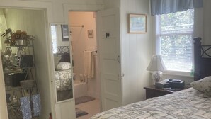 Une chambre à coucher comprenant un lit, un ventilateur de plafond, une fenêtre avec des stores et une porte donnant sur une salle de bain.