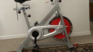 Un vélo d’appartement est placé dans une pièce dont le sol est recouvert d’un tapis à motifs et dont un des murs est uni.