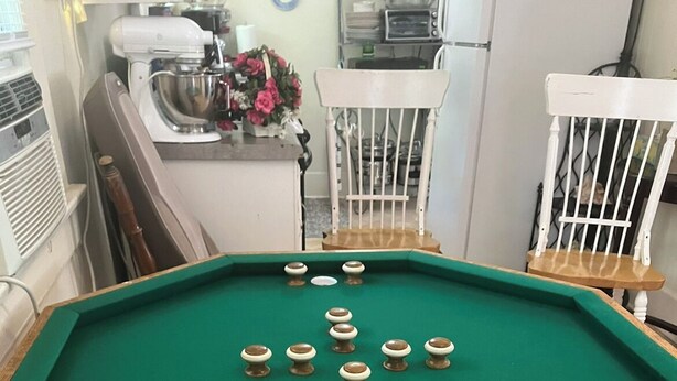 Une table de billard recouverte d’un drap vert, entourée de chaises en bois et d’une table blanche.