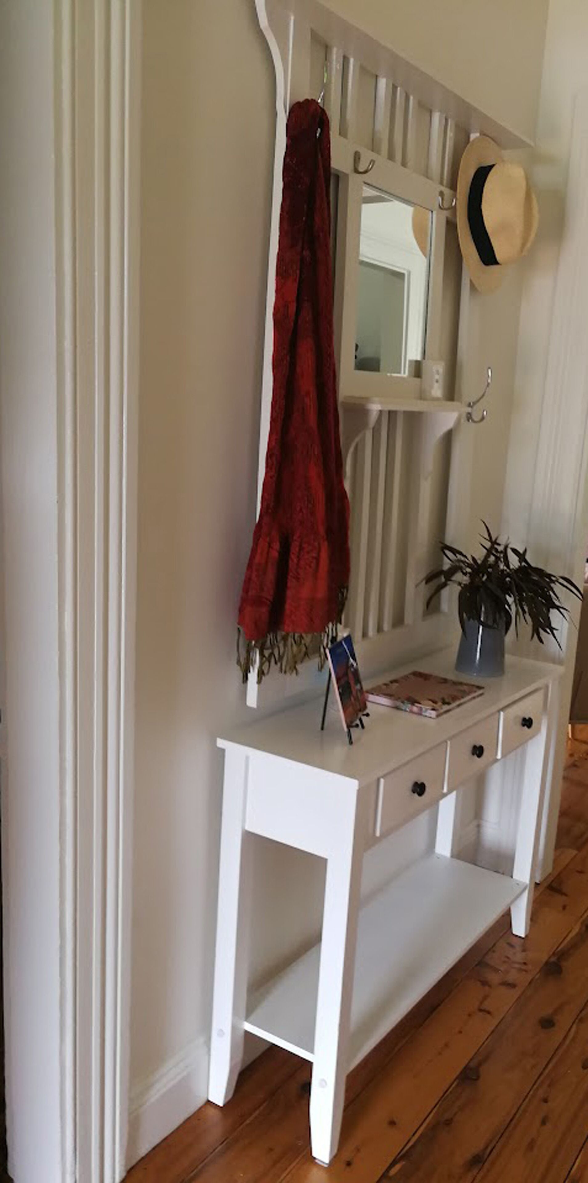Een witte console tafel met een spiegel erboven, een rode handdoek aan de muur, en een hoed aan een haak.