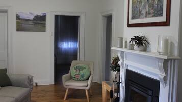 A living room with a fireplace, a sofa, a chair with a floral cushion, and two paintings on the wall.