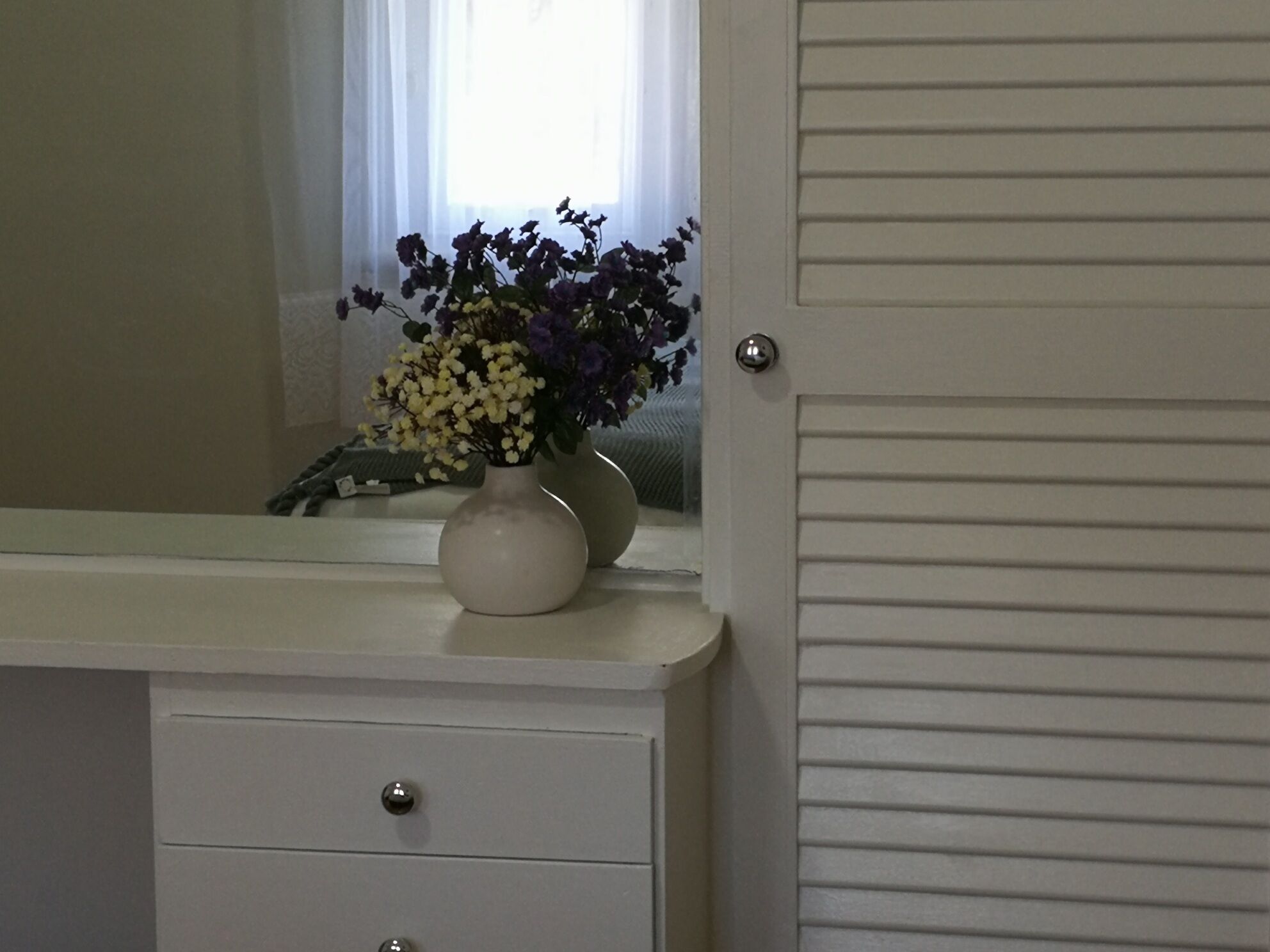 Ein Badezimmer-Waschtisch mit Spiegel, eine Vase mit Blumen und eine Tür mit Griff.