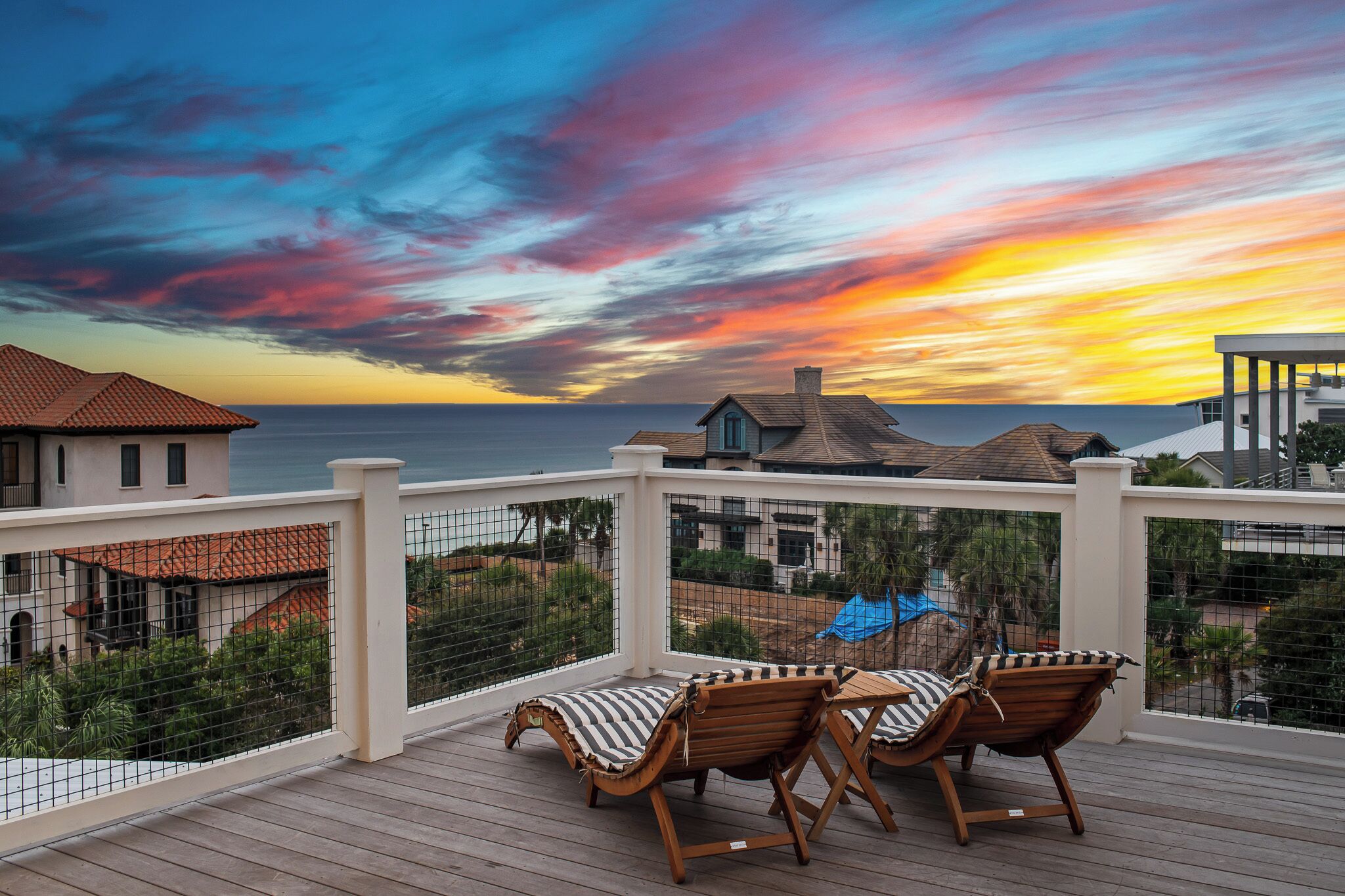 A wooden deck with two lounge chairs and a table, overlooking a coastal area with houses and a sunset.