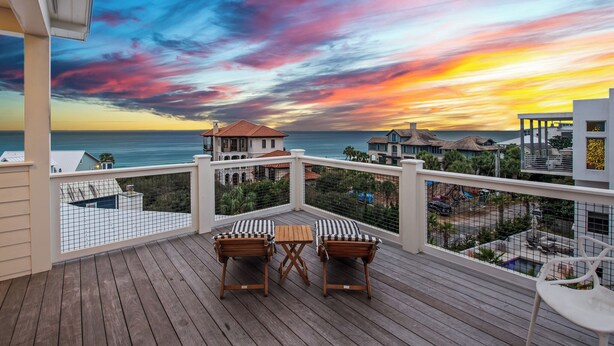A wooden deck with two chairs and a table, overlooking a coastal area with buildings and a sunset. - 12 Periwinkle Ln - Beach Blessings (Santa Rosa Beach)