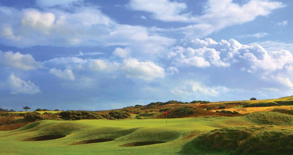 A golf course with rolling hills, a red flag marking the hole, and a clear sky.