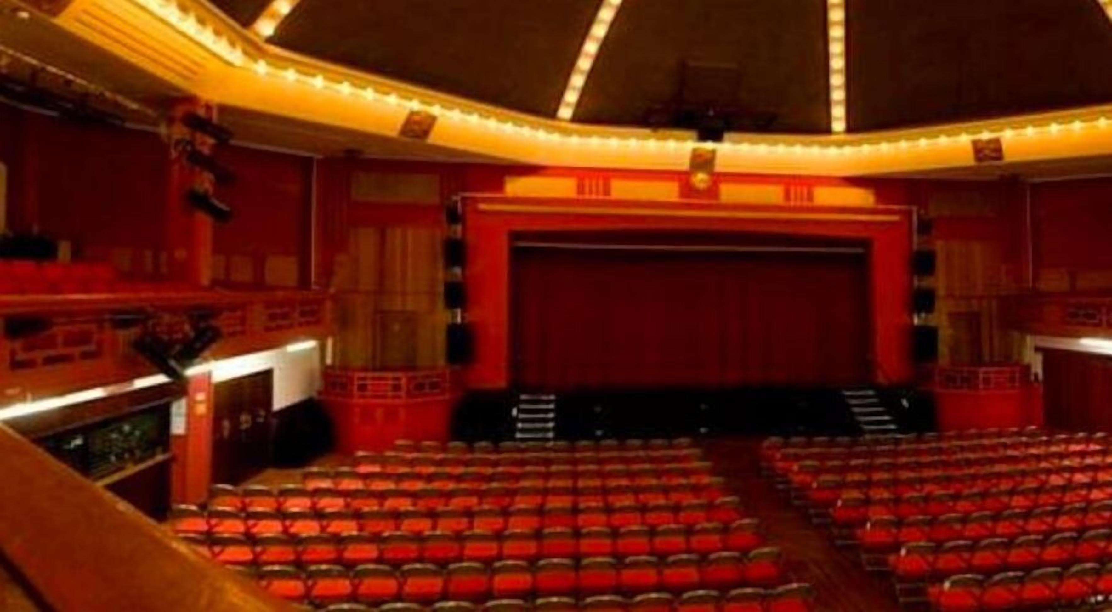En teatersal med rækker af røde sæder, en scene og en balkon.