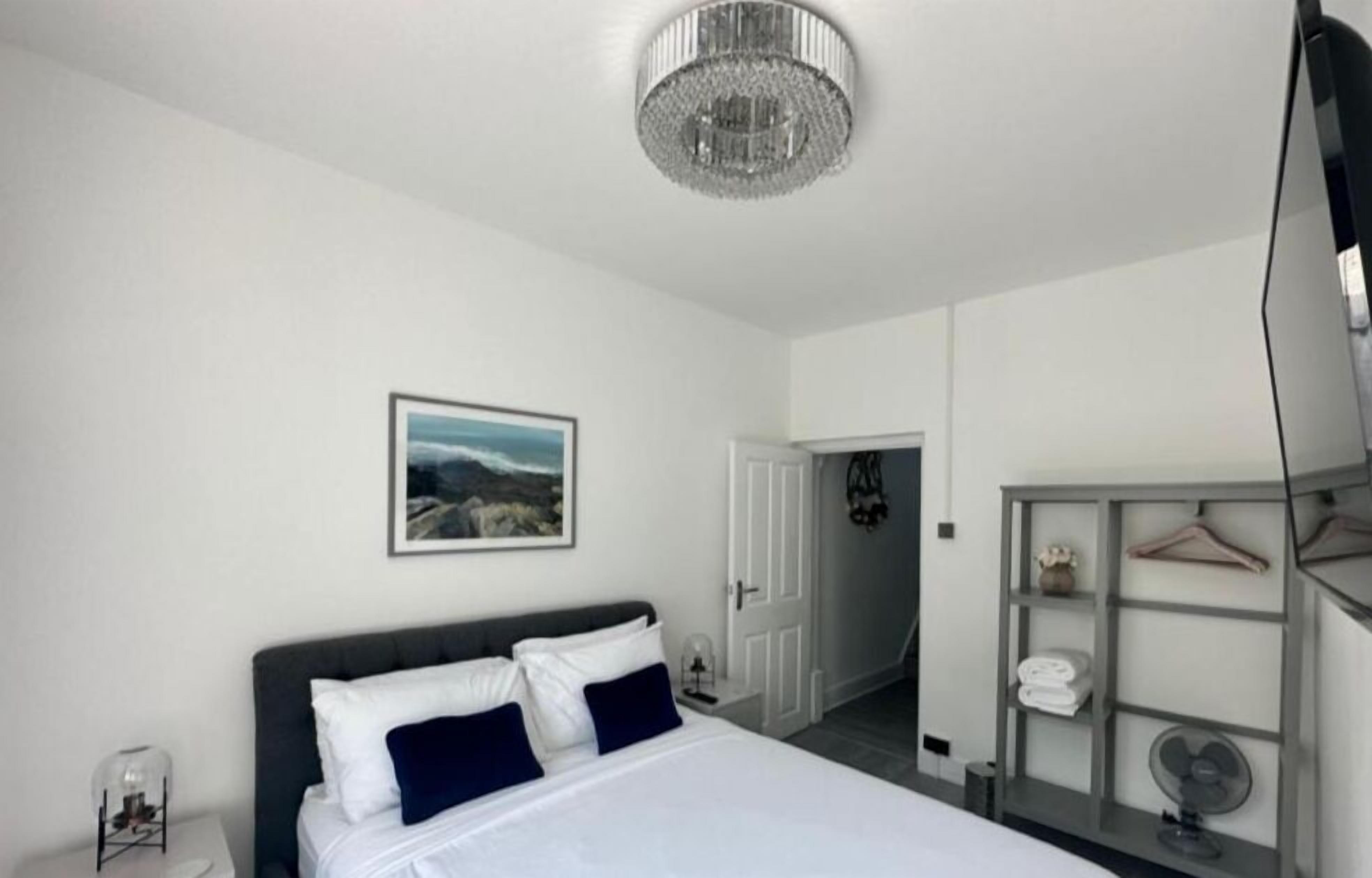 A bedroom with a bed, a chandelier, a framed picture, a wardrobe, and a door leading to another room.