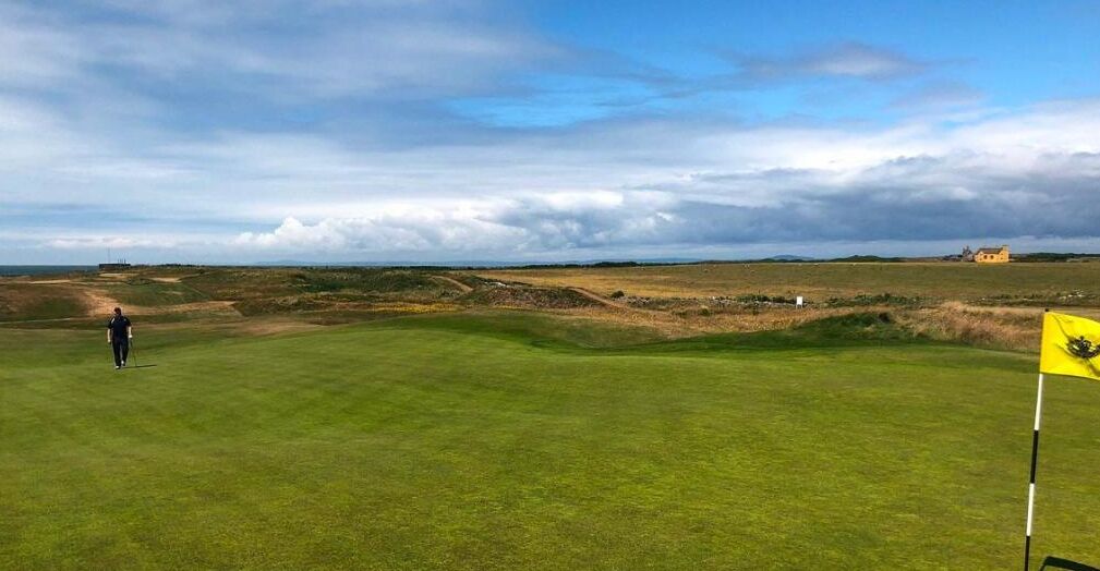 Un terrain de golf avec un drapeau, une personne qui marche et un bâtiment situé au loin.