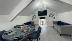 Ein Essbereich mit einem Glastisch, blauen Stühlen und einer Chandelier. Ein Sofa mit blauen Kissen und ein an der Wand befestigtes Fernsehgerät sind ebenfalls vorhanden.