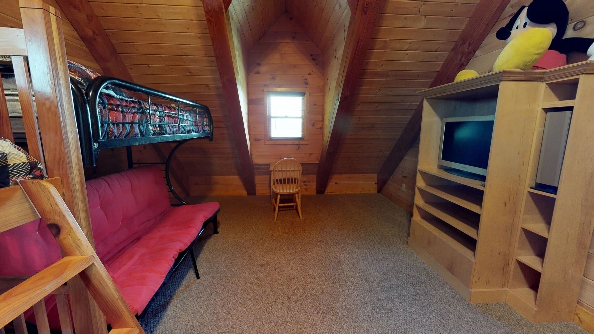 A cozy attic room with a red sofa, a small wooden chair, a television on a wooden stand, and a window letting in natural light.