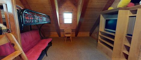 A cozy attic room with a red sofa, a small wooden chair, a television on a wooden stand, and a window letting in natural light.