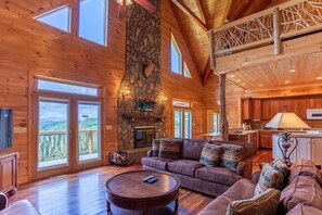 A spacious log cabin interior with a stone fireplace, wooden furniture, and large windows offering a view of the surrounding forest. - Family Lodge with Sunset Views (Elk Park)