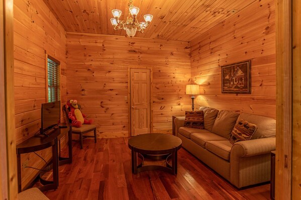 Un acogedor interior de cabaña de madera con piso de madera, un sofá, una mesita, un televisor y una araña.