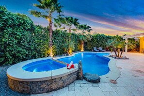 A swimming pool with a stone edge, surrounded by lush greenery and palm trees. - Flairbnb/Pool/Spa/Pool Table (Indio)
