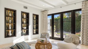 Una sala de estar moderna con un sofá blanco, una mesa de centro redonda de madera y cojines decorativos. Hay estantes montados en la pared con jarrones y un ventanal con cortinas.