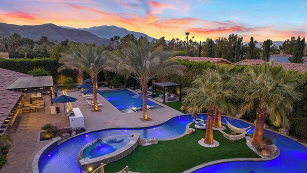 Un área de piscina con jardines, varias piscinas, palmeras y mobiliario exterior.