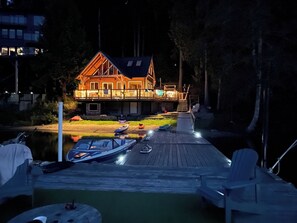 Un muelle de madera con barcos y sillas, una casa con terraza, y una masa de agua tranquila.