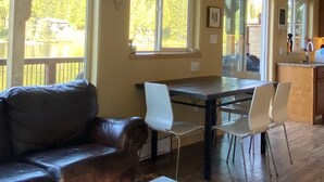 Een ruime kamer met houten plafond, een groot raam met uitzicht op een meer, een zithoek met een tafel en stoelen, en een gezellige zithoek met een bank en salontafel.