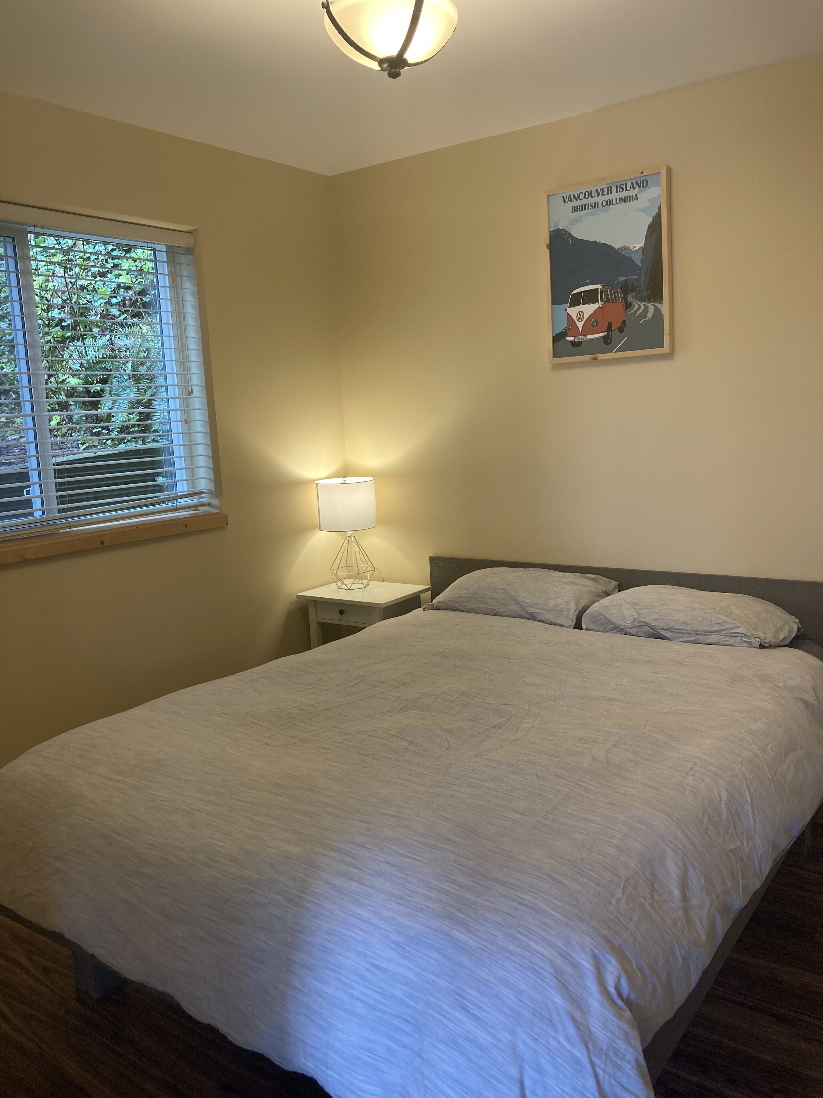 A bedroom with a bed, a nightstand, a lamp, a window with blinds, and a poster on the wall.