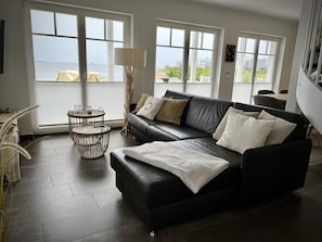 Sala de estar moderna com um sofá de couro preto, uma mesa de centro redonda de madeira e vista para o mar através de grandes janelas.