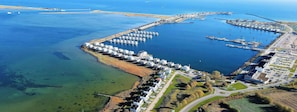 Vista aérea de un puerto deportivo costero con numerosos barcos amarrados, un puerto con un muelle largo y una zona residencial con casas y carreteras.
