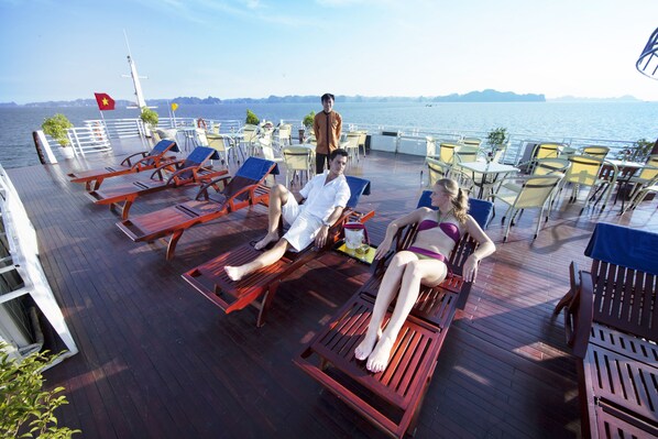 A deck with sun loungers, tables, and chairs, overlooking a body of water.