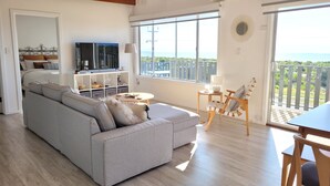 A living room with a grey sofa, a TV, a small table, and a chair. There is a balcony with a view of the sea. - Mint by the Sea - Esplanade location, sea views! Free WiFi! Off Peak Special! (Tiddy Widdy)