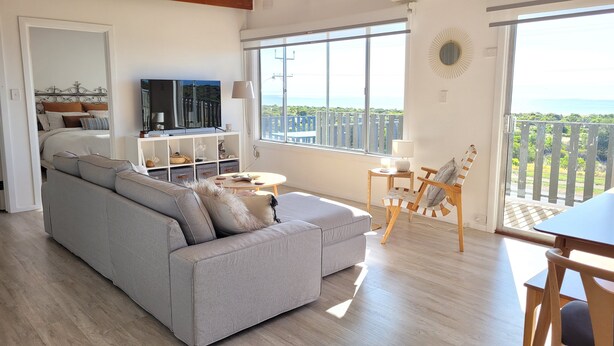 Una sala de estar con un sofá gris, un televisor, una mesita y una silla. Hay un balcón con vistas al mar.
