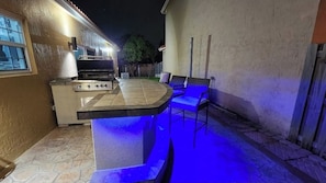An outdoor kitchen area with a grill, bar seating, and blue ambient lighting.