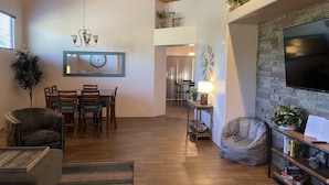 Ein geräumiges Wohnzimmer mit Holzbalkendecke, einem Essbereich mit Holztisch und -stühlen, einem großen Flachbildfernseher an einer Steinwand und einer gemütlichen Sitzecke mit Sitzsack und Bücherregalen.