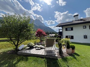Un edificio bianco con una terrazza in legno, un albero fiorito di colore rosso e una montagna sullo sfondo.