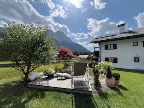 A white building with a wooden deck, a red flowering tree, and a mountain in the background. - Frühholz—The Vacation Home, 5-Star "Wank" Apartment in Grainau & Shared Garden (Grainau)