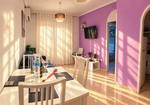 A well-lit room with a dining table, chairs, a television mounted on the wall, and a window with curtains.