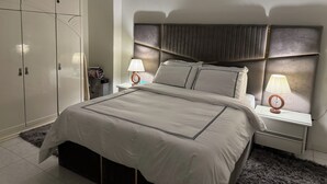 A neatly arranged hotel room with a large bed, two bedside tables with lamps, a wardrobe, and a small desk with a chair. - Nile View Apartment - Sleeps 4 & Balcony (Cairo)