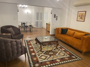 Ein Wohnzimmer mit Sofa, Sessel, Glastisch, gemustertem Teppich, einer Essecke mit Stühlen und Tisch sowie einer Küche im Hintergrund.