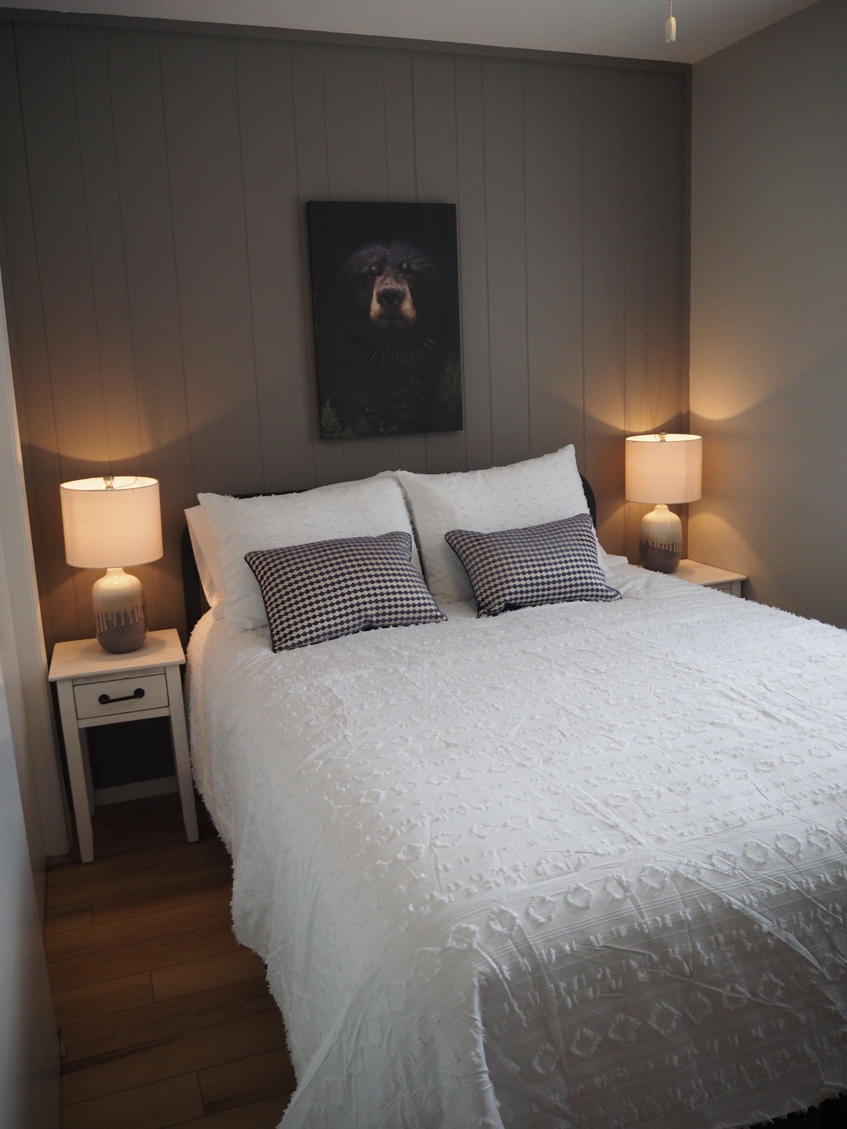 A neatly made bed with white linens and patterned pillows, flanked by two bedside tables with lamps, in a room with wooden flooring and a framed picture on the wall.