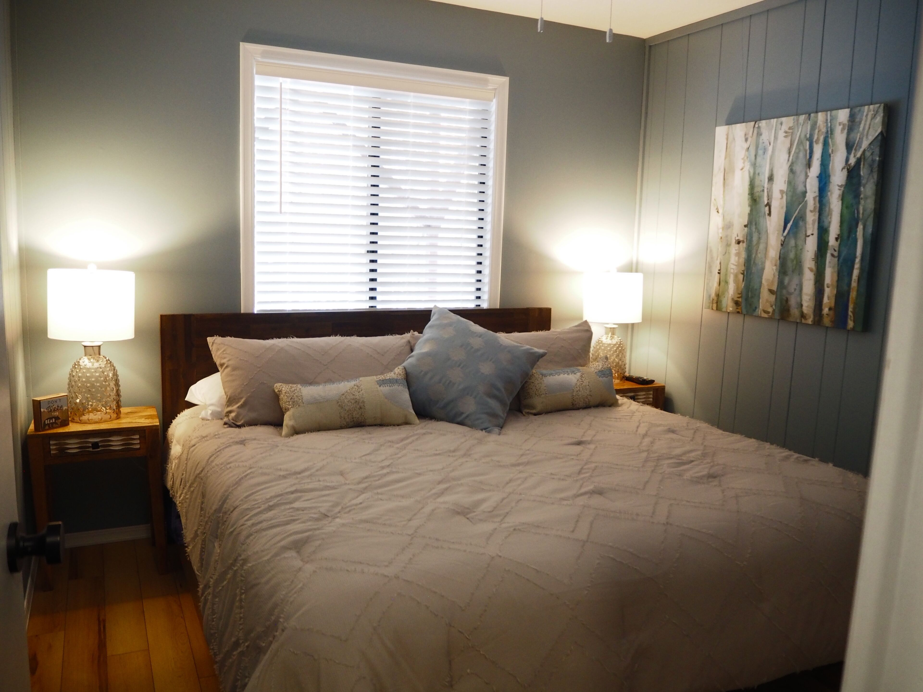 A bedroom with a large bed, two bedside tables with lamps, a window with blinds, and a wall-mounted painting.
