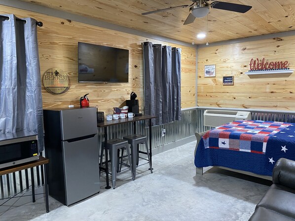 A compact hotel room with a kitchenette, a bed with a patriotic quilt, a television, and a ceiling fan.