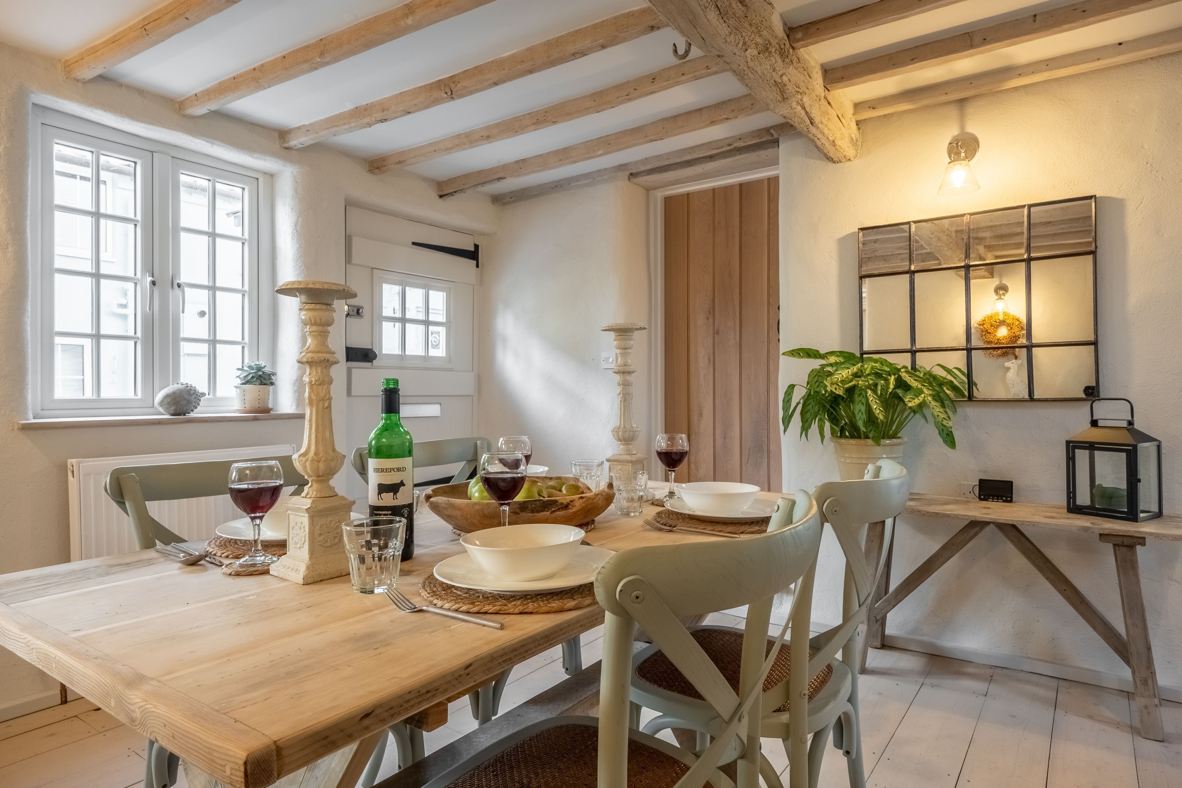 Un coin repas avec une table en bois dressée pour quatre personnes, entourée de chaises blanches. On peut voir un plafond en bois avec des poutres apparentes, et un luminaire fixé au mur se trouve au-dessus de la table.