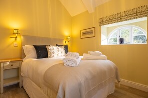 A neatly made bed with white linens and a folded towel, a bedside lamp, and a window with a view of greenery. - The Coach House (R), Ringstead, Norfolk (Ringstead)
