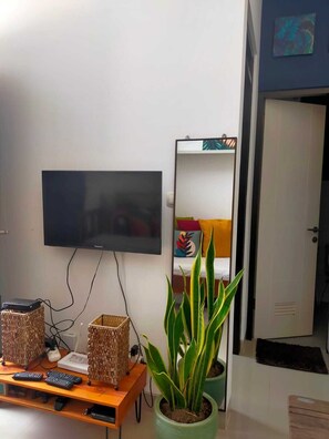 A living room with a flat-screen TV mounted on the wall, a wooden coffee table with remote controls, a potted plant, and a mirror reflecting a colorful pillow and a bed. - The Valley Guesthouse (Malang)