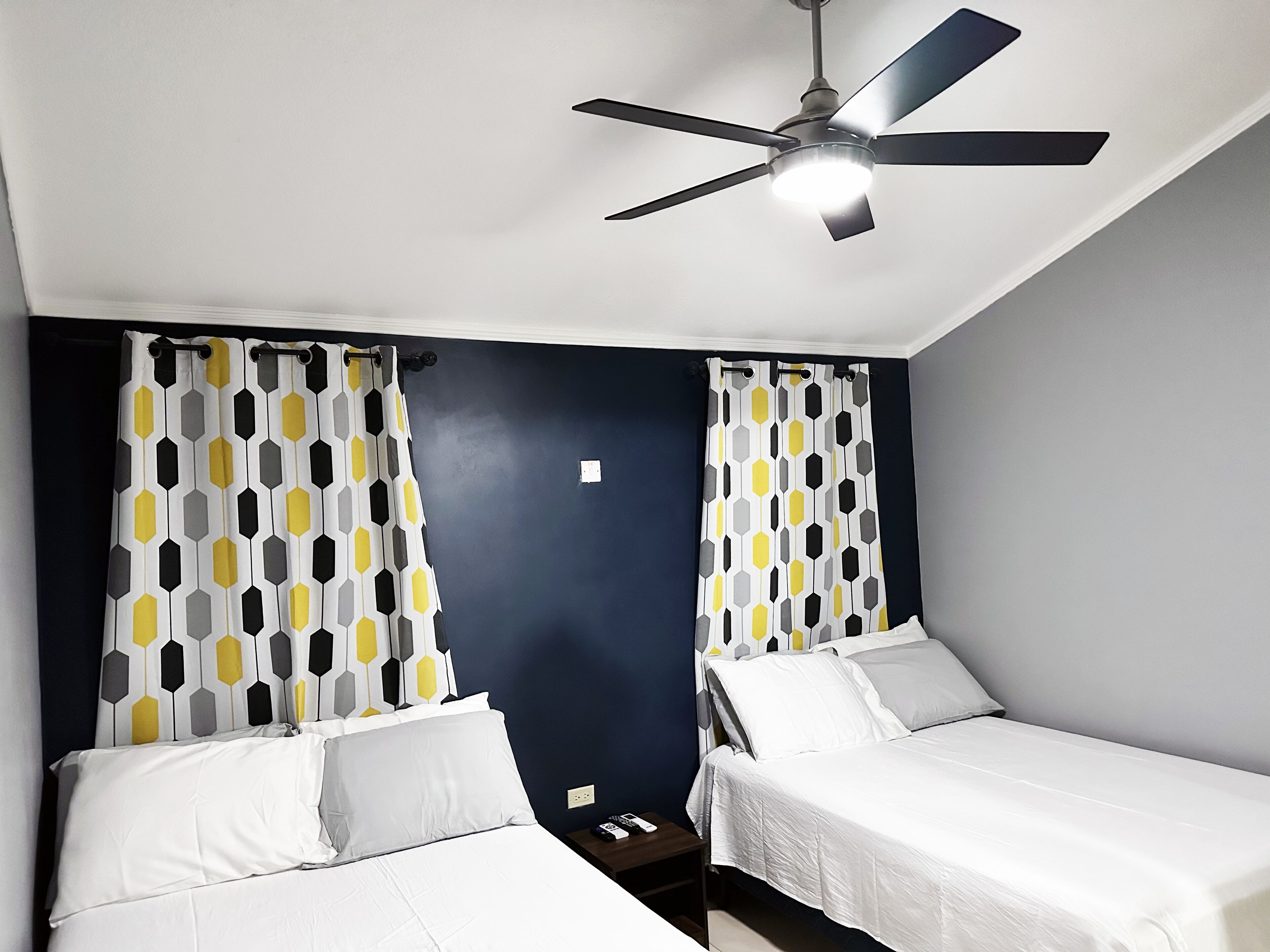 A room with two single beds, a ceiling fan, and curtains with a geometric pattern.